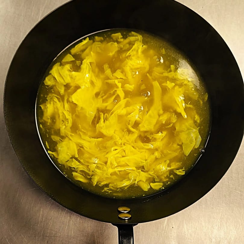 A black pan filled with yellow egg drop soup, bubbling on a stove top.