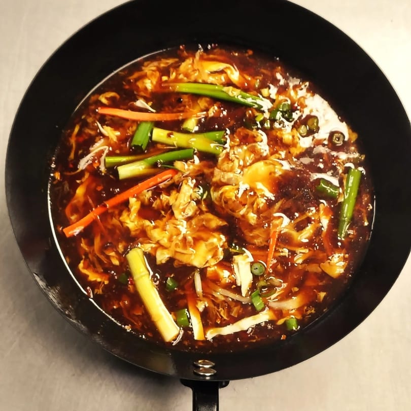 A close-up of Chinese hot and sour soup with scallions, carrots, and egg in a black bowl.