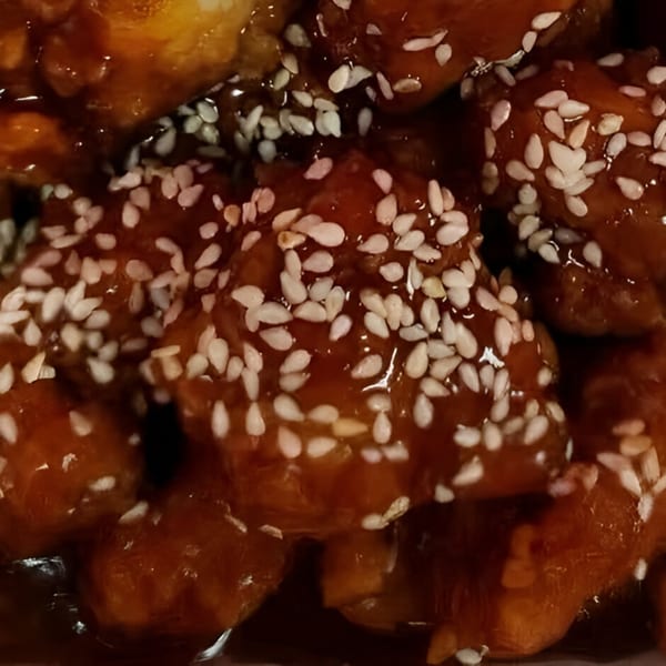Close-up of sesame chicken pieces coated in a glossy, dark orange sauce with white sesame seeds.