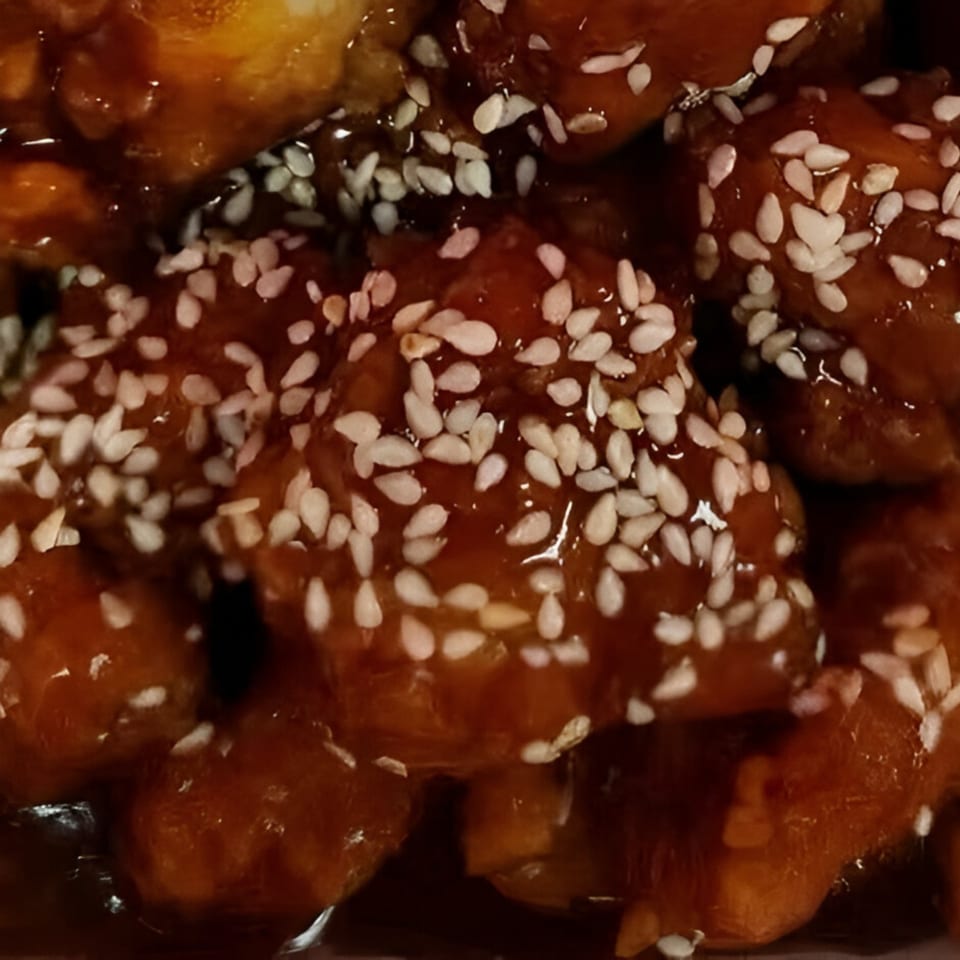Close-up of sesame chicken pieces coated in a glossy, dark orange sauce with white sesame seeds.